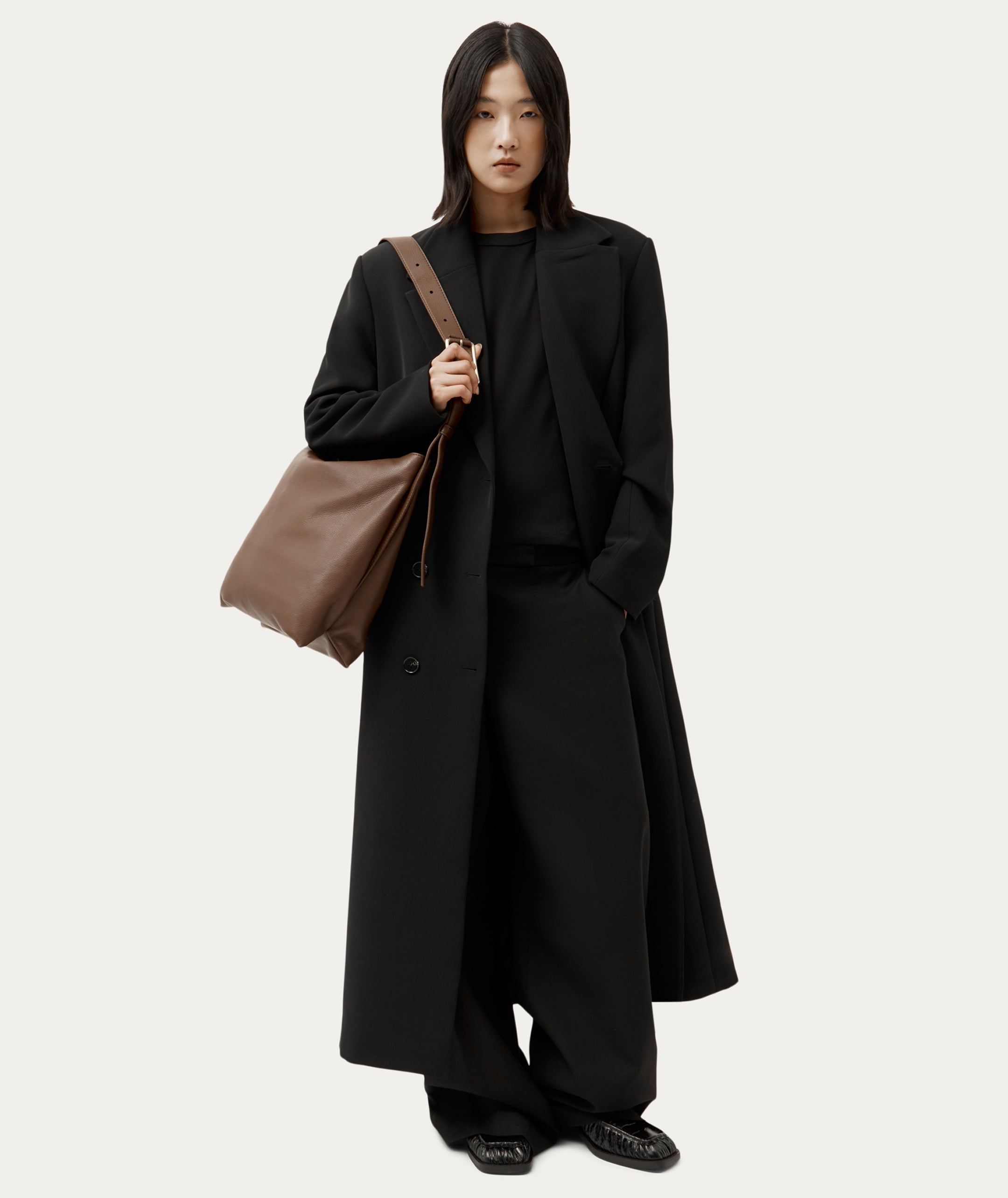 A model wearing a long black coat, wide-leg trousers, and black shoes, carrying a large brown leather shoulder bag against a light gray studio background.