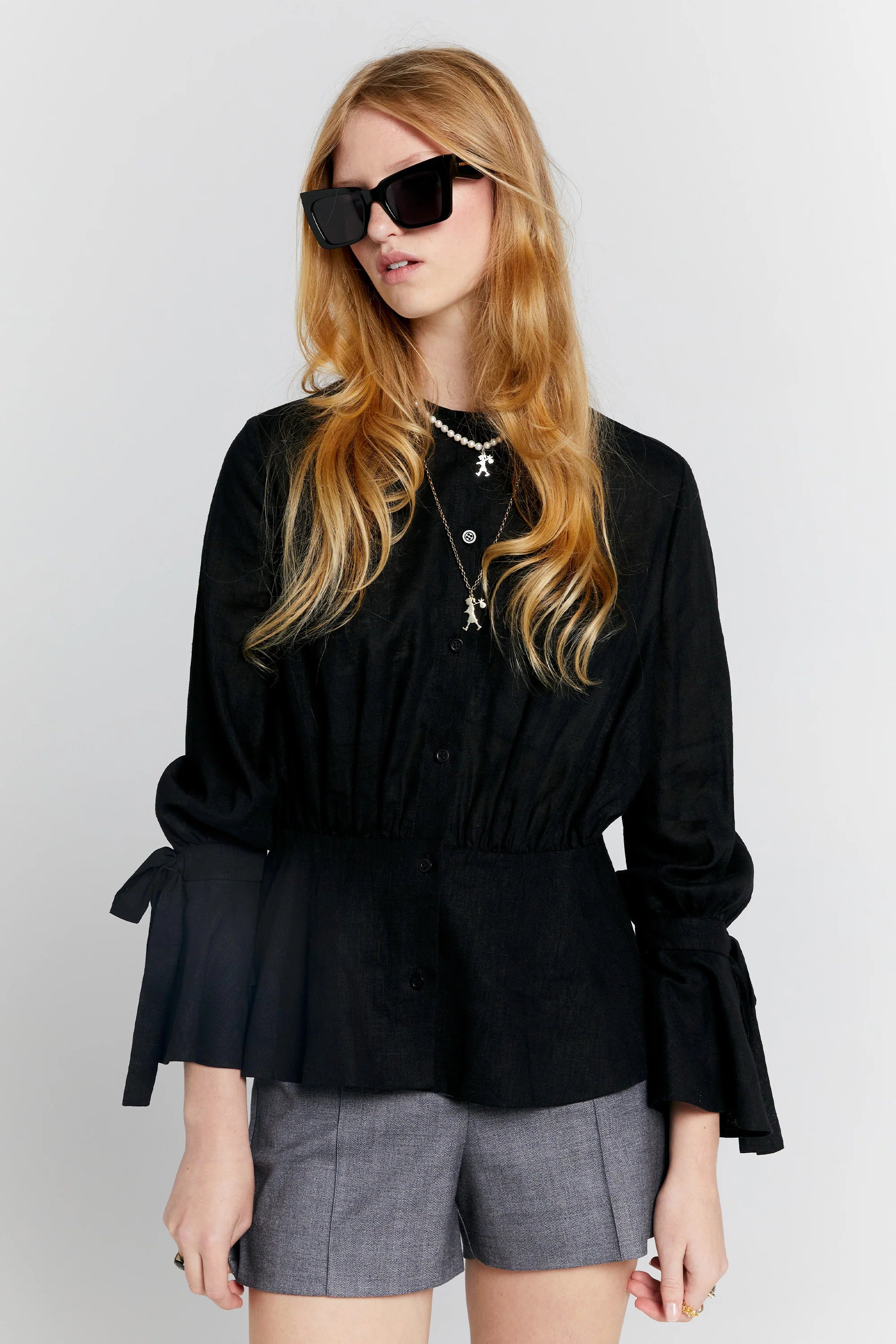 Female model with long blonde hair wears dark sunglasses and a long sleeve black top that has flared sleeves with ties at the wrist, no collar, and flares from the waist with a button stand through the centre front. 