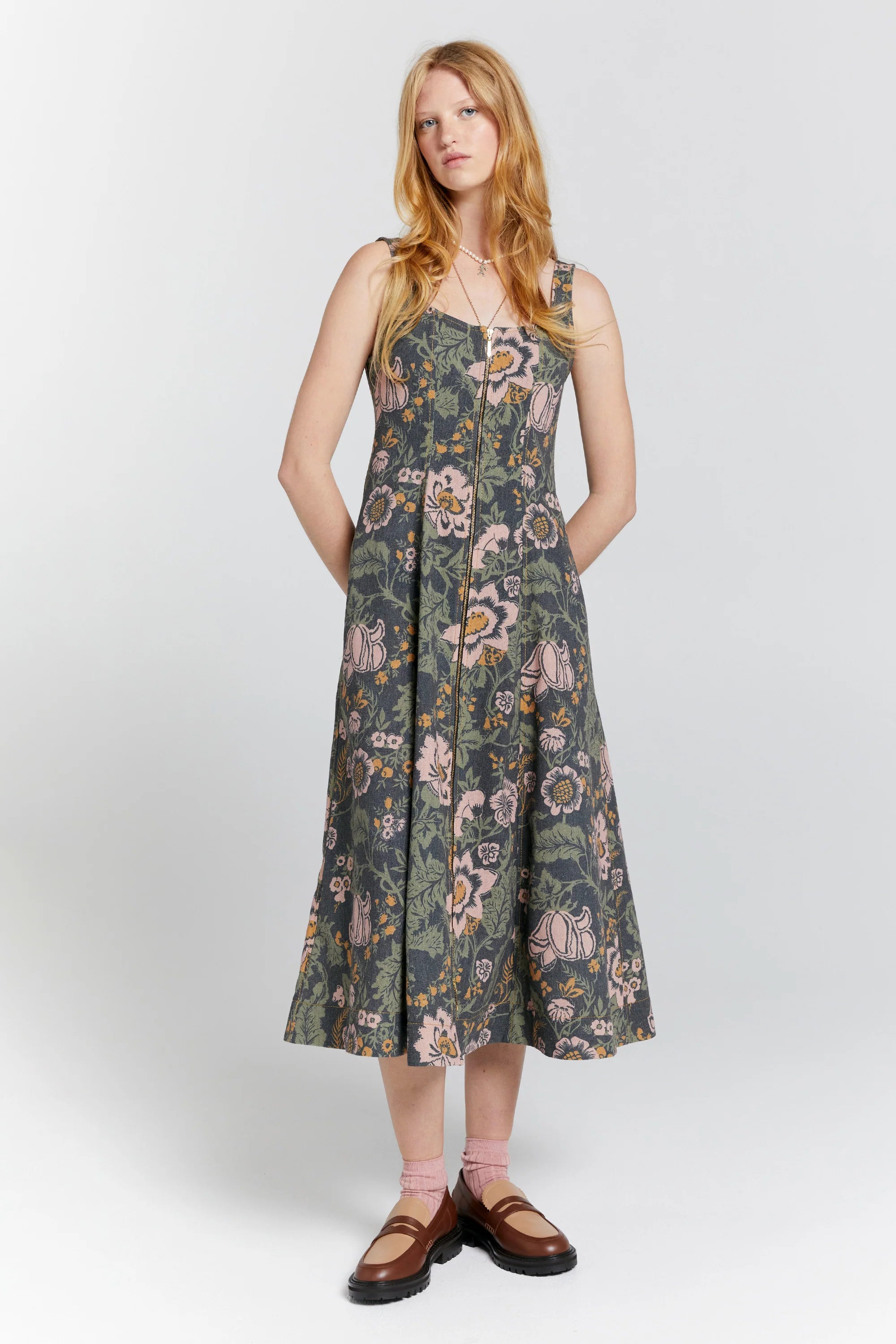 A female model with long blonde hair stand front on with hands behind her back. Wearing a floral print dress that is fitted through the bodice and flares from the waist, features a gold zip with arrow pull tabs right through the front. Dress is sleeveless with thick singlet style straps.