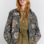 Female model with long blonde hair wears retro style large sunglasses with gradient lenses. An olive green shirt and floral printed linen blend bomber style jacket with gold zip.