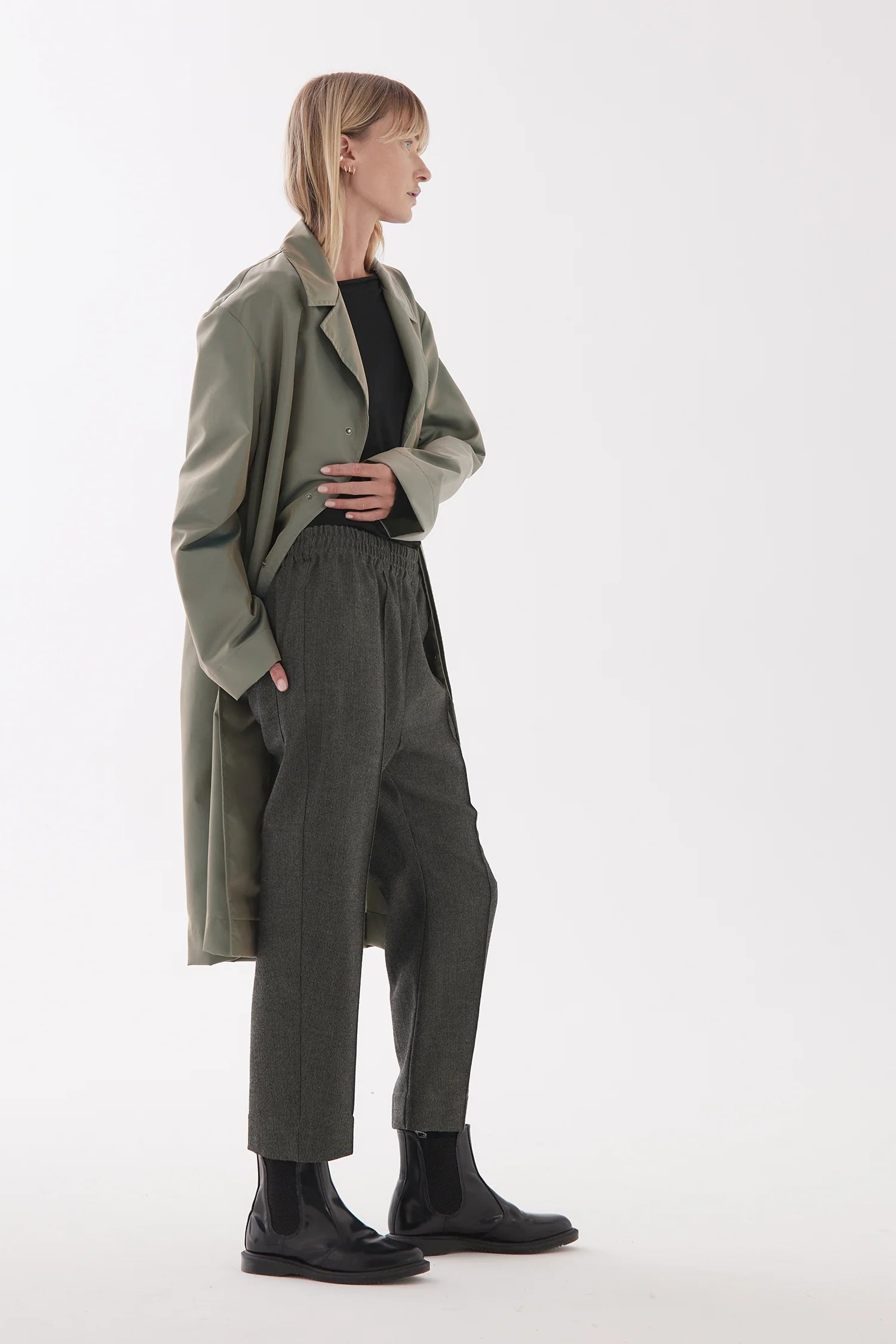 Female model with light hair wears a long light green coat with elasticated drop crotch small herringbone patterned pants that fall to the ankle. Model stands to the side and is wearing black boots