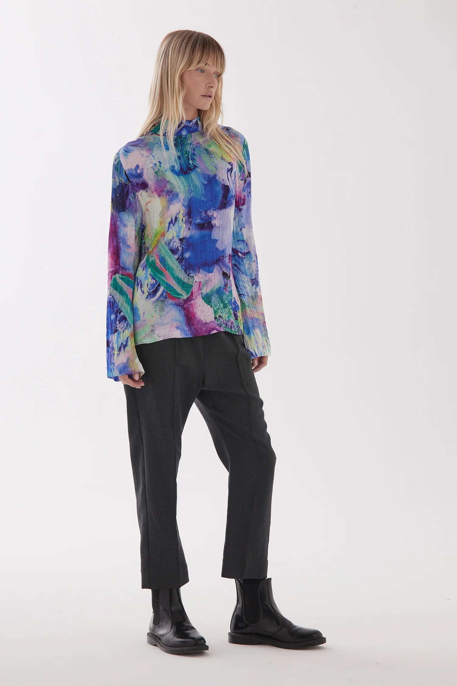 Female model with long light hair stands facing to the side but body straight on and wears a multicoloured high neck top with long sleeves and elasticated pull on relaxed crotch pants with pintuck seams on the front legs. Pants fall to the ankle and mode wears black ankle boots