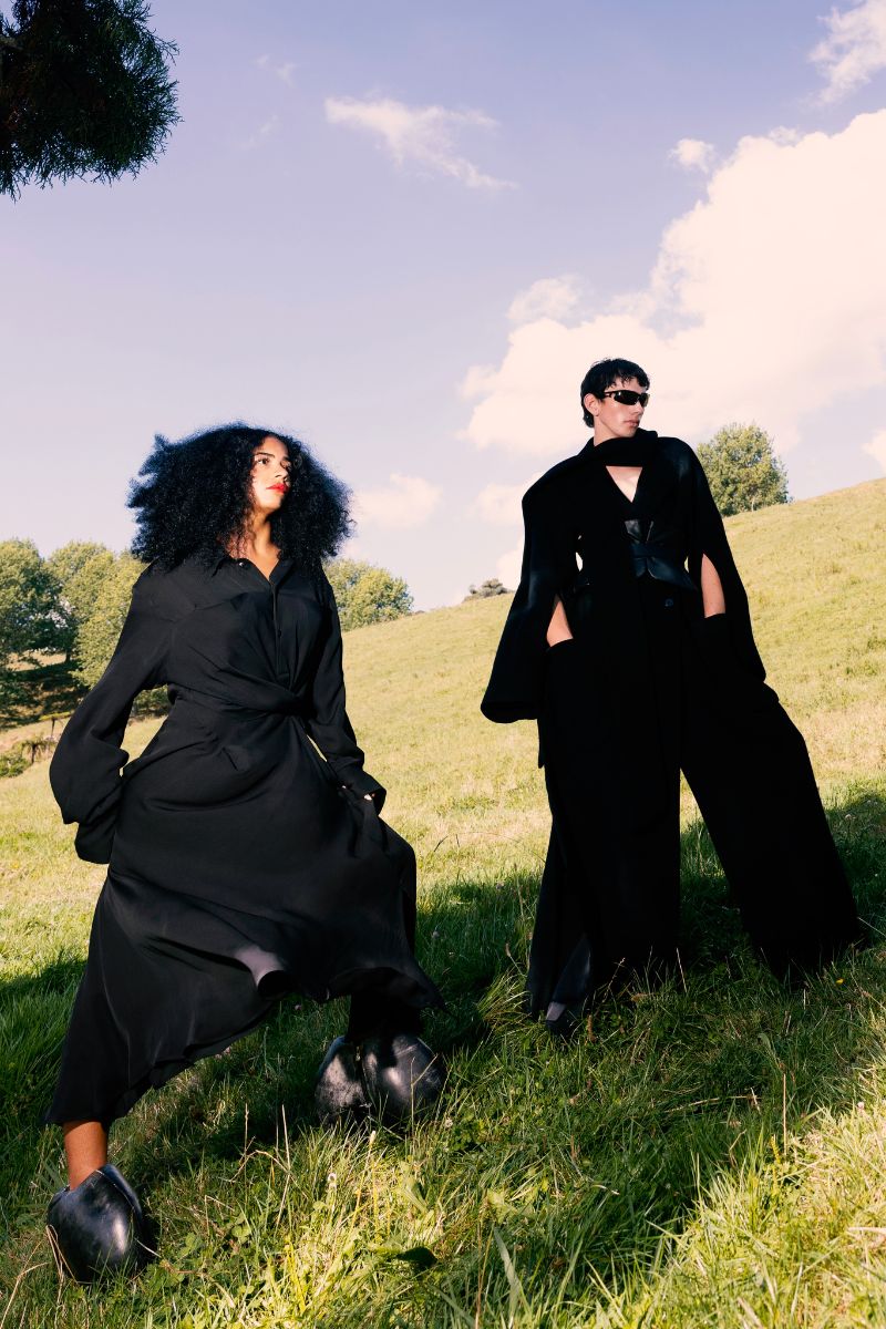 Two people in black outfits standing on a grassy field with trees and blue sky in the background.
